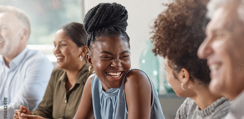 woman with her braids pulled up into a bun in a blue sleeveless shirt, sitting down surrounded by co-workers, she is in the middle. She is smiling at the coworker on her left.