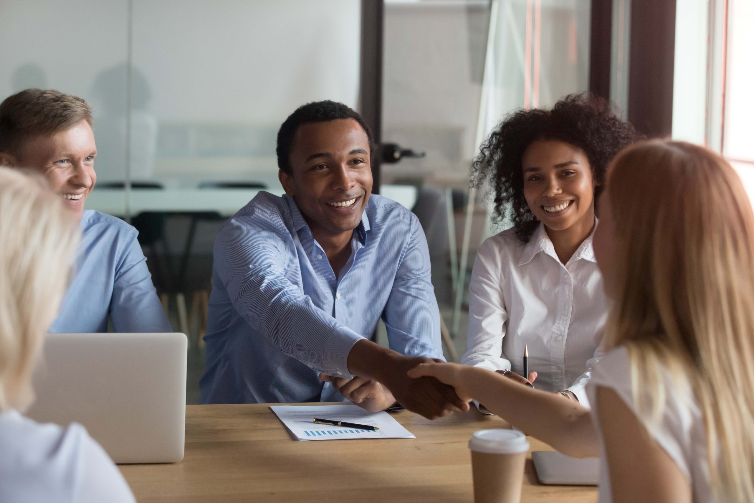 Successful diverse teammates sitting at boardroom, company head shaking hands with millennial specialist woman businesspeople ready to start conference discuss financial report planning future goals.